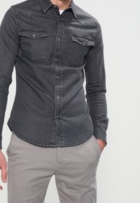 Man som bär en åtsittande mörkgrå jeansskjorta med två bröstfickor och ljusgrå byxor, med ena handen i fickan.