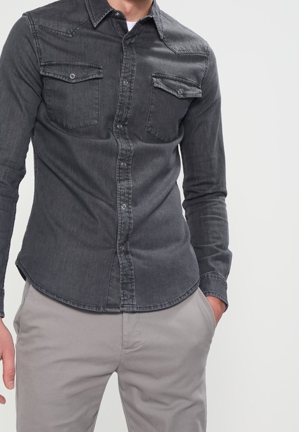 Man som bär en åtsittande mörkgrå jeansskjorta med två bröstfickor och ljusgrå byxor, med ena handen i fickan.