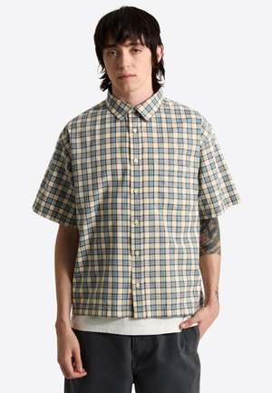 Jeune homme aux cheveux foncés portant une chemise à manches courtes à carreaux beige, bleu et vert, par-dessus un t-shirt blanc et un pantalon noir.