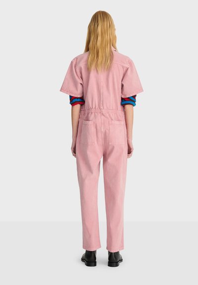 Personne blonde debout, de dos, portant une combinaison rose à manches courtes par-dessus un vêtement à manches longues rayé bleu et rouge, ainsi que des bottines noires.
