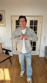Young man standing indoors, smiling, wearing light gray hoodie over white t-shirt, blue jeans, and sneakers, buttoning his hoodie.