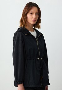Zwarte lichtgewicht jas met een capuchon, elastische taille en gouden hardware. Voorzien van lange mouwen met elastische manchetten en een rits aan de voorkant.