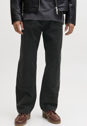 Uomo che indossa pantaloni neri a gamba larga, scarpe marroni in pelle con lacci, giacca di pelle nera e camicia scura, in piedi contro uno sfondo neutro.