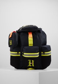 Zaino navy con accenti gialli riflettenti, multiple tasche frontali, dettagli in rete e una piccola pochette staccabile. Dotato di maniglia superiore e spallacci regolabili.