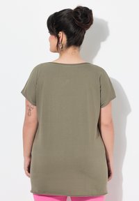 Tunique olive verte ample avec des manches courtes, texture douce, ourlet arrondi et design minimal. Présente un tatouage sur le bras gauche.
