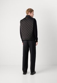 Black quilted vest with a high collar, featuring diamond patterns. Worn over a black long-sleeve shirt and paired with dark pants and shoes.