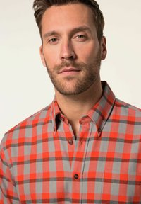Camisa de cuadros rojo y gris con botones y cuello, que presenta detalles en negro, dos botones y un bolsillo de parche en el lado izquierdo.