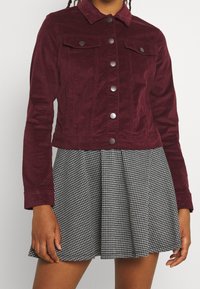 Veste en velours côtelé bordeaux avec boutons-pression et deux poches poitrine, associée à une jupe plissée à carreaux noir et blanc.