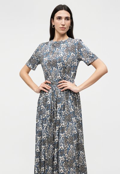 Femme aux longs cheveux foncés portant une robe bleue à manches courtes avec un motif floral, debout les mains sur les hanches devant un fond uni.
