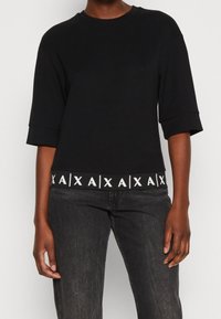 Černé tričko s krátkým rukávem a kulatým výstřihem, se bílým logem na lemu. Měkká tkanina s uvolněným střihem.