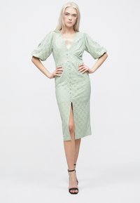 Myntgrønn kjole med perforert design, dyp V-hals, puffede ermer og sidesliss. Tøylekstrukturen virker lett og pustende.