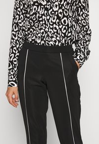 Femme portant un pantalon noir à rayures blanches verticales et une chemise à manches longues à imprimé léopard noir et blanc, une main dans la poche.