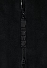 Gros plan d'une fermeture éclair noire sur un tissu noir avec une sangle verticale attachée au curseur de la fermeture, présentant des lettres texturées sur la sangle.