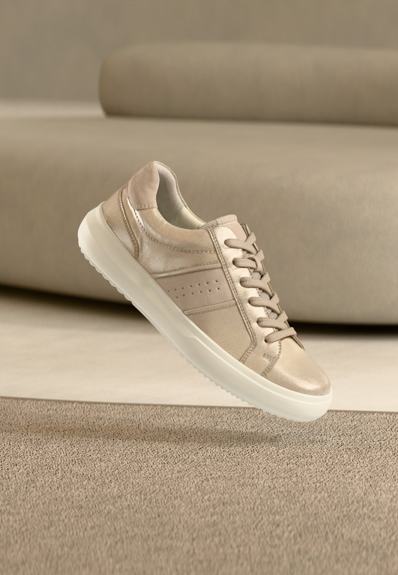 Μπεζ αθλητικό παπούτσι με κορδόνια και λευκή σόλα που αιωρείται πάνω από υφασμένο μπεζ χαλί, ουδέτερος καναπές θολωμέν ος στο φόντο.