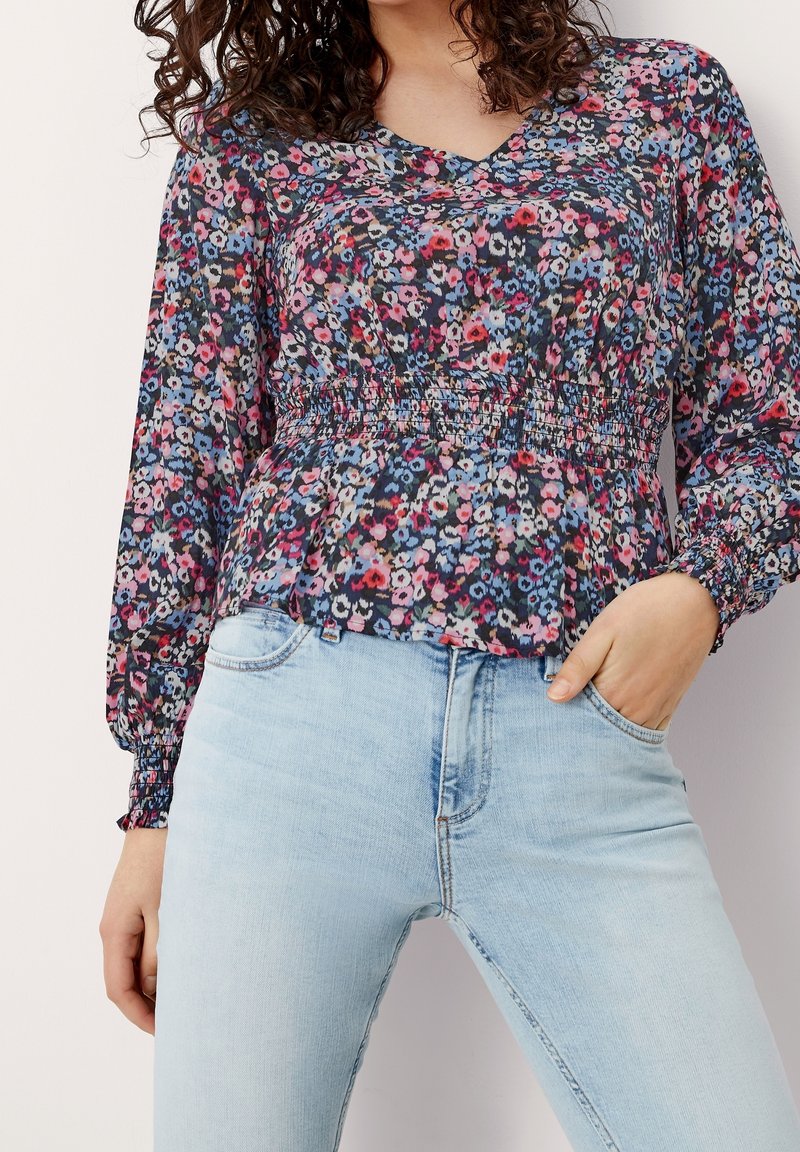 Blusa floreale su sfondo scuro con fiori rosa, blu e bianchi. Tessuto leggero, vita arricciata e maniche lunghe. Abbinata a jeans in denim chiaro.