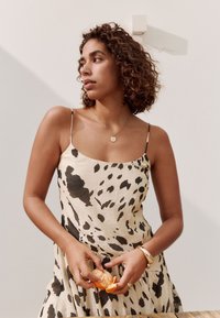 Vrouw met krullend haar in een zwart-wit patroon jurk die een sinaasappel schilt, draagt gouden sieraden, staande tegen een witte muur.