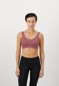 Kvinne med kort mørkt hår iført en mauve sports-BH og høyt liv svarte leggings, stående mot en ensfarget hvit bakgrunn.