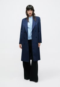 Žena s tmavými vlasmi v dlhom námorníckomodrom dvojradovom kabáte, svetlom modrej blúzke a širokých čiernych nohaviciach stojaca pred jednofarebným pozadím.