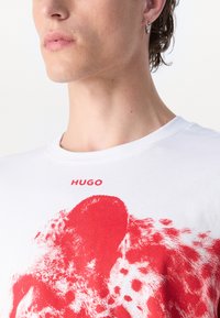 White T-shirt featuring a red abstract design. "HUGO" is printed above the graphic. The fabric appears smooth with a classic crew neck.