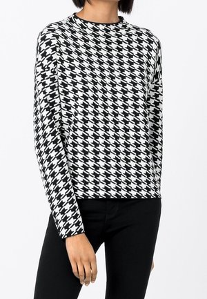 Woman wearing black and white houndstooth long-sleeve sweater and black pants, standing against a plain white background.