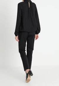 Femme portant un chemisier noir à manches longues avec un col à boutons, un pantalon noir ajusté et des escarpins noirs, se tenant devant un fond blanc.