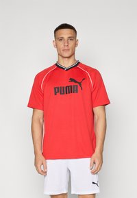 Rød sportsdrakt laget av lettveid materiale med svarte detaljer og v-hals. Har en fremtredende svart "PUMA"-logo på fronten.