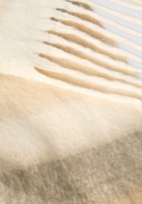 Coperta in lana con una gradazione di colori crema, beige e taupe, caratterizzata da una texture morbida e bordi frangiati per un tocco di dettaglio in più.