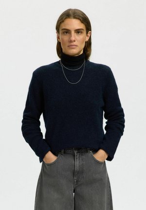 Femme aux cheveux bruns porte un col roulé noir sous un pull bleu foncé, un jean gris et un fin collier en argent, mains dans les poches.