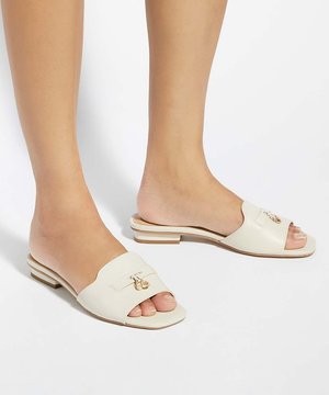 Pieds portant des sandales crème ouvertes à l'avant, à enfiler, avec de petites décorations dorées et des talons bas empilés sur un fond blanc.