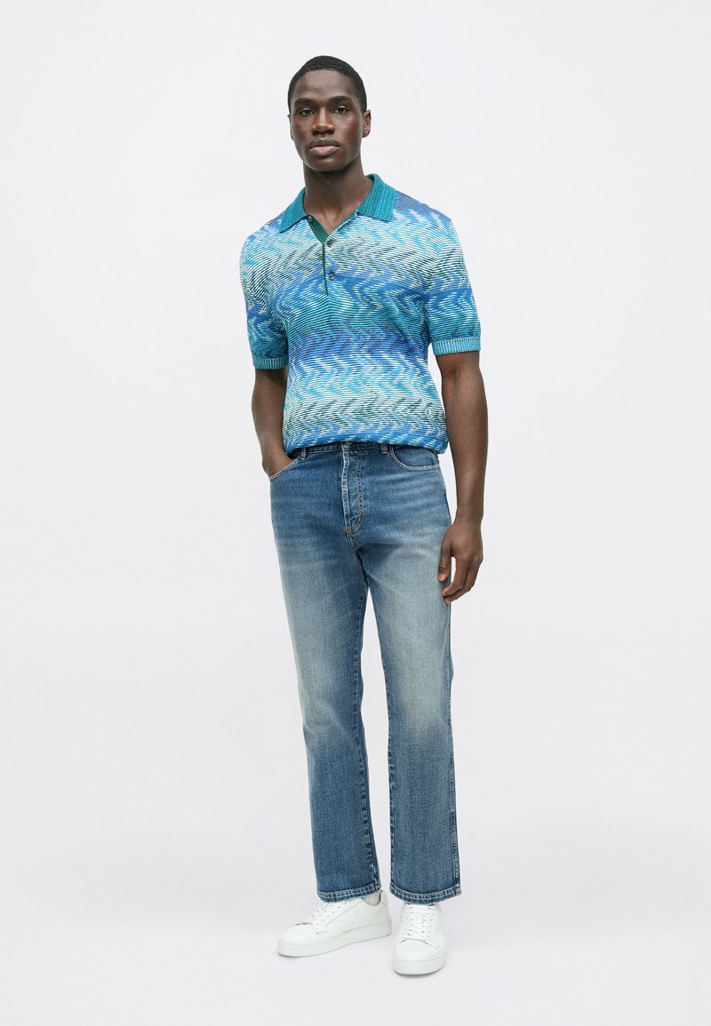 Polo bleu à motifs avec col, associé à un jean bleu clair à jambes droites et des baskets blanches. Texture lisse et coupe moderne.