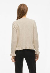 Bež pulover s volančki na robu in manšetah, mehak na otip, dolgi rokavi in sproščen kroj. Brez vidne zapore. Nosimo ga čez črne hlače.
