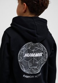 Zwarte hoodie met een ronde witte grafische print met golvende lijnen en de tekst "HUMMEL". De stof lijkt textuur te hebben en casual te zijn.