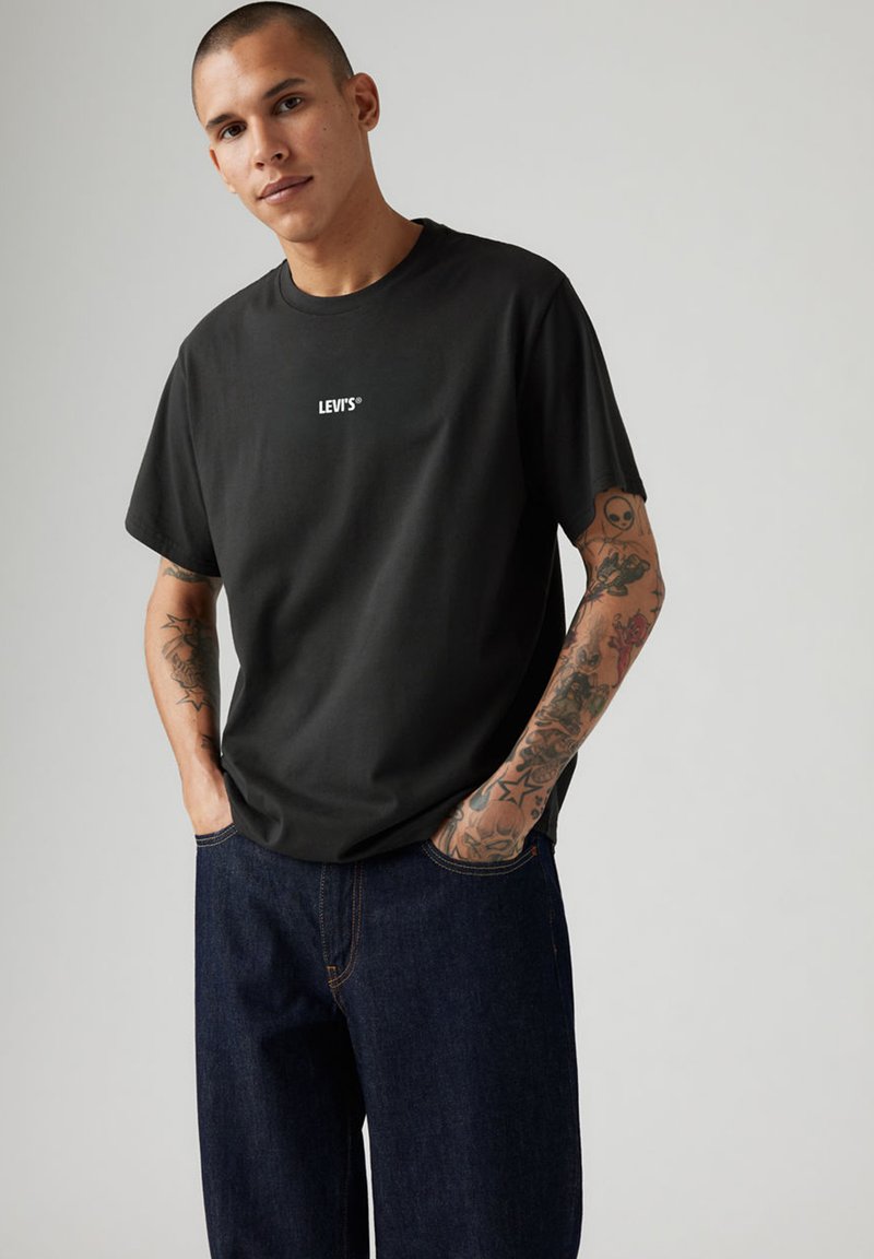 Jeune homme tatoué portant un t-shirt noir Levi’s et un jean bleu foncé, debout les mains dans les poches devant un fond uni.