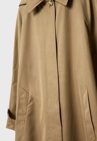 Trench-coat beige avec boutons avant dissimulés, deux poches inclinées, un col et un détail de sangle sur le poignet de la manche.