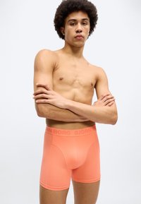 Ανοιχτό πορτοκαλί ανδρικά εσώρουχα boxer με ομαλή υφή, με λογότυπο στη μέση. Η εφαρμογή είναι στενή και ο σχεδιασμός χωρίς σχέδια ή επιπλέον διακοσμήσεις.