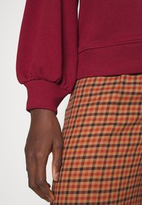 Sudadera burdeos con puños de manga larga abullonados, combinada con una falda de patrón a cuadros en tonos marrón y naranja. Detalle de tejido texturizado visible.