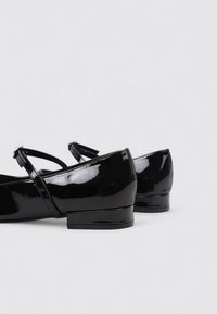 Bailarinas de charol negro con punta redonda, tacón bajo de bloque y correa en el tobillo. Textura lisa con acabado brillante.