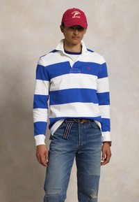 Tricou de rugby cu dungi albastre și albe, cu guler, asortat cu blugi deschis la culoare, deteriorați, și o șapcă roșie cu text alb.