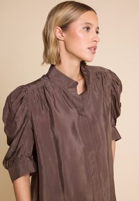 Brun satengbluse med puffede korte ærmer, mandarin krave og en glat tekstur. Har en knapfri frontplade for et strømlinet design.
