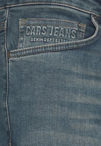 Blue denim jeans feature a textured fabric with a faded finish. The back pocket has a stitched label reading "CARS JEANS."