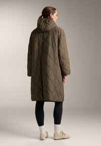 Olijfgroene gewatteerde lange jas met een capuchon, voorzien van een ruitpatroon. Het model draagt zwarte leggings en crèmewitte schoenen met witte sokken.