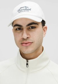 Balta beisbolo kepurė su mėlynu siuvinėtu tekstu "Susitiksime prie aikštelės" ir "lululemon įsteigta 1998". Dėvima su aukštu kaklu, užtraukiama kreminės spalvos striuke.