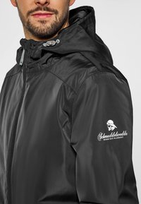 Chaqueta negra impermeable con capucha, que cuenta con un cierre frontal y un logo en la manga izquierda. Textura suave y diseño ligero.