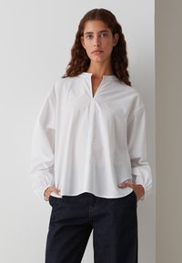 Weißes Langarmhemd mit gespaltenem Kragen und gerafften Manschetten, mit einem geraden Saum. Kombiniert mit dunkelblauen Jeans.