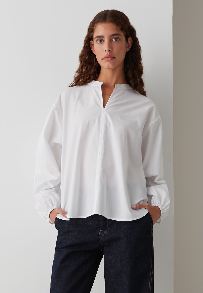 Weißes Langarmhemd mit gespaltenem Kragen und gerafften Manschetten, mit einem geraden Saum. Kombiniert mit dunkelblauen Jeans.