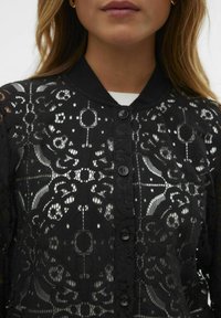 Blouse en dentelle noire présentant des motifs floraux complexes, un col et des boutons noirs sur le devant, avec une texture douce et transparente.