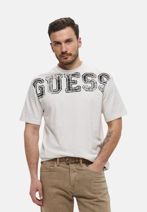 Uomo con capelli corti e scuri che indossa una maglietta bianca con testo nero "GUESS" e pantaloni beige, in piedi con una mano in tasca.