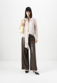 Frau in transparenter, gemusterter Bluse mit Halsband, weit geschnittenen braunen Lederhosen, schwarzen High Heels und einer blassen gelben Handtasche über einer Schulter.