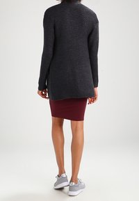 Mujer vestida con un cárdigan de punto oscuro, falda burdeos ajustada y zapatillas grises de pie con la espalda a la cámara.