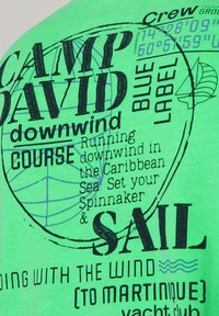 T-shirt verde in cotone con testo e grafiche stampate nere, inclusi elementi marittimi e coordinate geografiche, che conferisce un design casual.